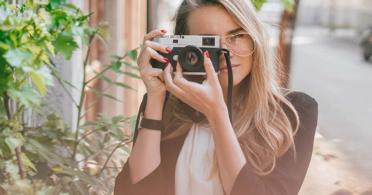 Bir kadın beyaz bir arka planın önünde durmakta ve sağ elinde bir dijital fotoğraf makinesi tutmaktadır. Sol kolu bükülmüş, kamerayı göğsüne yakın tutuyor. Odaklanmış bir ifadeyle objektife doğru bakıyor. Siyah bir gömlek giymiş gibi görünüyor ve koyu kahverengi saçları düşük bir at kuyruğu şeklinde arkaya toplanmış. Fotoğraf makinesinin kendisi, büyük bir objektifi ve siyah gövdesi olan gelişmiş bir dijital SLR. Objektif arka plandan gelen ışığı yansıtarak ona parlak, metalik bir görünüm veriyor. Ön planda, yaprakları beyaz arka plan tarafından aydınlatılan küçük, yeşil bir bitki görülüyor. Kadının pozu ve ifadesi fotoğraf çekmenin ortasında olduğunu gösteriyor.