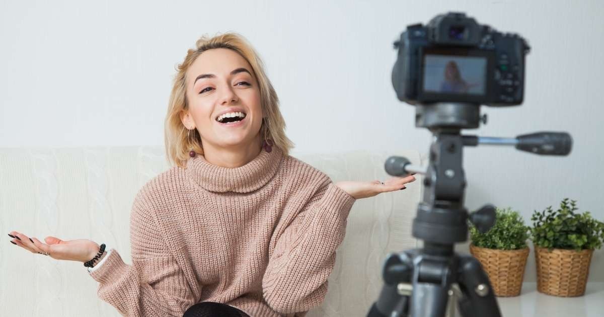 Bir kadın elinde fotoğraf makinesiyle kanepede oturuyor. Yüzünde parlak bir gülümseme var ve sanki fotoğraf çekmeye hazırmış gibi ellerini uzatmış. Yan tarafında sepet içinde bir bitki ve bileğinde siyah bir elastik bant görülüyor. Fotoğraf makinesinin bir ekranı var ve onu tutan el yana doğru uzanıyor. Kadının boynuna yakın çekim yapılmış ve gülüyor gibi görünüyor. Kamera, kanepenin ve bitkinin sıcak, canlı renkleriyle büyük bir kontrast oluşturuyor. Bu görüntü bir neşe ve rahatlama anını tasvir ederken, teknolojinin önemini ve zaman içinde bir anı yakalamayı da ihmal etmiyor.