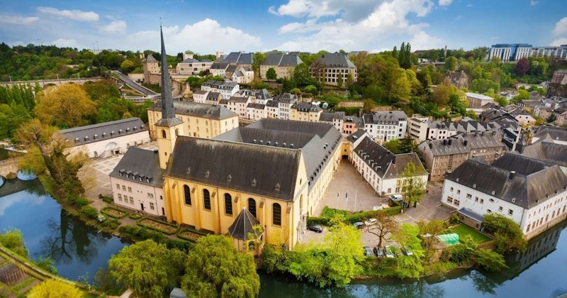 Bu, beyaz bir çan kulesi ve görüntünün ortasından geçen dolambaçlı bir nehri olan büyük bir bina. Gökyüzü masmavi ve bulutlarla kaplı; binanın bazıları açık olan çok sayıda penceresi var. Binanın yanına park edilmiş bir araba ve binanın üzerinde siyah bir çatı var. Nehrin kenarında küçük bir ağaç grubu ve binadan uzağa giden bir patika var. Arka planda ağaçlar ve bir bina ile nehrin manzarası çok güzel.
