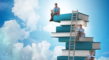 Kitapların üzerinde oturan iki adam görülüyor. Soldaki adam gri bir gömlek ve mavi bir kot pantolon giymiş ve aşağıya doğru bakıyor. Sağdaki adam ise mavi bir gömlek giymiş ve kameraya doğru bakıyor. Her iki adam da bacak bacak üstüne atmış ve rahatlamış görünüyor. Arkalarında, mavi gökyüzü ve beyaz bulutların olduğu büyük bir pencere görülüyor. Görüntünün sağ tarafında bir kişi merdivenlerde oturmuş kitap okumaktadır. Görüntünün sol tarafında bir adam elinde dizüstü bilgisayarıyla bir sandalyede oturuyor. Daha solda, bir adam merdivende otururken kitap okurken görülüyor. Resmin sol alt köşesinde bir kişi dizüstü bilgisayar kullanırken görülüyor. Son olarak, görüntünün sağ alt köşesinde bir grup insan bir merdivenin üzerinde otururken görülüyor.