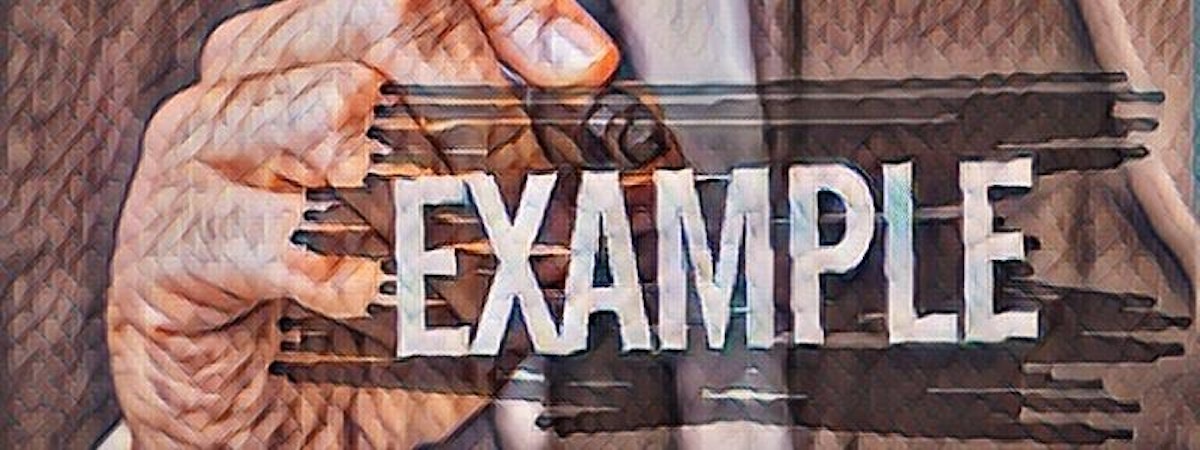 This image shows a close-up of a right hand holding a long, cylindrical cigar. The hand has a light tan complexion and is wearing a black dress shirt with the cuff rolled up. The cigar is brown in color with a few lighter shades in the wrapper. It is slightly curved and looks like it has been recently lit. Smoke is slowly rising from the end. The background is blurry and out of focus. The focus is on the hand and the cigar. The setting appears to be indoors.