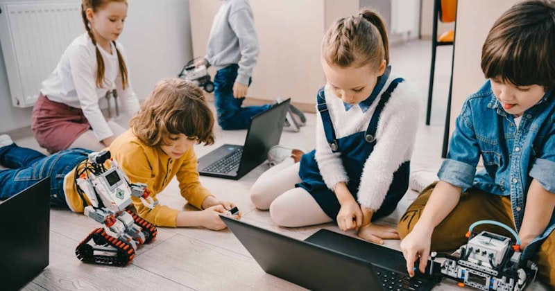 Bir grup çocuk bir odada oturmuş, dizüstü bilgisayarla oynuyor. Atkuyruklu bir kız çocuğu ön planda bir masanın üzerinde otururken, yaramaz bir ifadeye sahip genç bir oğlan çocuğu arka planda dilini çıkarıyor. Çerçevenin ortasında bir çocuk bir robotla etkileşime giriyor, bir eliyle ona dokunurken diğer eliyle bir dizüstü bilgisayarı manipüle ediyor. Dizüstü bilgisayarın yakın çekiminde, tuşların üzerinde gezinen bir kişinin eli görülüyor. Yerde açık duran bir dizüstü bilgisayar, oyun oynamanın keyfini çıkaran bir grup çocuk tarafından unutulmuş gibi görünüyor.