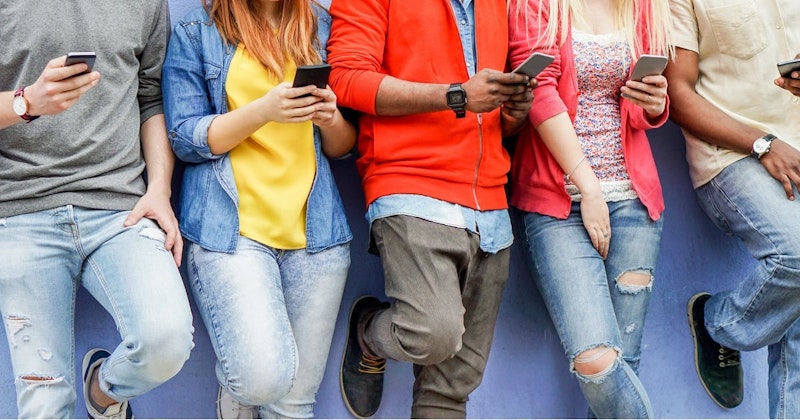 Bir grup insan birbirine yakın durmakta ve çoğunluğu kameraya bakmaktadır. En soldaki kişi sarı bir gömlek ve mavi bir kot pantolon giyerken, sağındaki kişi yırtık bir kot pantolon üzerine kırmızı bir hırka giyiyor. Ortadaki kişi kırmızı bir kazak ve gri bir pantolon giymiş ve elinde bir cep telefonu tutuyor. Sağdaki kişi duvara yaslanmış ve turuncu bir ceket giymiş, elinde de bir telefon var. Daha sağda sarı bir gömlek ve mavi kot pantolon giyen bir kişi var ve bir bacağı eğilmiş. Görüntüdeki tüm insanlar farklı yönlere bakmaktadır.