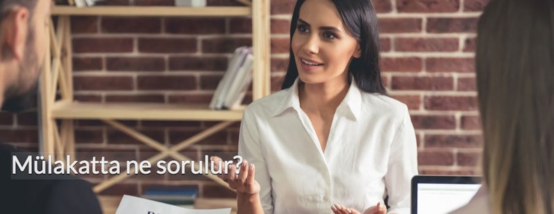 Kadrajın ortasında beyaz bir gömlek giymiş bir kadın durmaktadır. Saçları topuz yapılmış ve gözleri dümdüz karşıya odaklanmış. Ayaklarını hafifçe açmış, kolları iki yanında duruyor. Yüz ifadesi nötrdür ve izleyiciye hafif bir gülümsemeyle bakmaktadır. Gömleği düz ve kıvrılmamış ve kumaş hafifçe kırışmış. Arka plan biraz bulanık ama kadın net bir şekilde odaklanmış. Solda silik bir erkek yüzü görüntüsü, sağda bir merdiven ve arka planda bir kişinin eli ve saçı var. Genel olarak, görüntü sakin bir konsantrasyon anını yakalıyor.