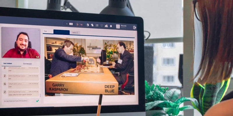 Kırmızı gömlekli bir adam ve takım elbiseli bir adam bilgisayar ekranında satranç oynamaktadır. Takım elbiseli adam yüzünde odaklanmış bir ifadeyle masanın üzerine eğilmiş. Arka planda, bulanık bir görüntüye sahip yeşil bir bitki var. Ayrıca, bir kutunun yakın çekimi ve bir kalemin yakın çekimi vardır. Son olarak, görüntünün köşesinde bir binanın bulanık bir görüntüsü var. Tüm bu unsurlar bir araya gelerek satranç oyununun yoğunluğunu anlatan bir sahne oluşturuyor.