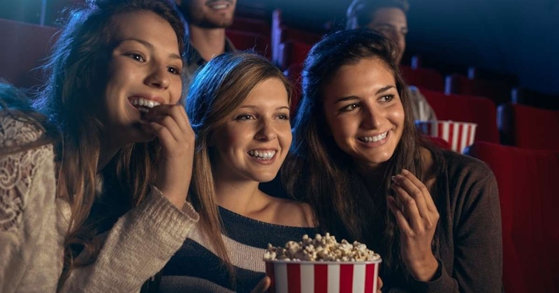 Bir grup mutlu kadın, patlamış mısır dolu kırmızı beyaz çizgili büyük bir kovanın etrafında toplanmış. Bir kadın elini yüzüne götürmüş genişçe gülümserken, diğer kadınlar yemek yerken gülüyorlar. Ön planda, yakın plan bir kadının gülümseyen yüzünü ortaya çıkarıyor, gözleri sevinçle parıldıyor. Arka planda bir kişinin yüzünün bulanık bir görüntüsü görülüyor. Hepsi birlikte eğlenceli ve rahat bir anın tadını çıkarıyor gibi görünüyor.