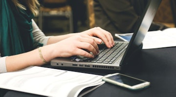 Bu görüntü bir dizüstü bilgisayarda yazı yazan bir kişiyi göstermektedir. Dizüstü bilgisayar çerçevenin büyük kısmını kaplıyor ve kişi önünde oturmuş, elleri klavyenin üzerinde. Kişi mavi bir gömlek giyiyor ve açık kahverengi saçları var. Dizüstü bilgisayar açık ve ekran aydınlatılmış. Dizüstü bilgisayarın beyaz bir klavyesi ve siyah bir trackpad'i vardır. Kişinin parmakları klavyede yazmaktadır ve rahat bir yazma pozisyonundadır. Arka planda bir kadının yüzünün yakın çekimi, bir kitabın yakın çekimi ve bir bileziğin yakın çekimi var. Çerçevenin sağ tarafında ise bir cep telefonunun yakın çekimi yer alıyor. Çerçevedeki aydınlatma sıcak ve doğal. Hepsi bir arada, bu görüntü bir dizüstü bilgisayarda yazı yazan birinin bir anını yakalıyor ve arka plandaki unsurlar ek bağlam ve ayrıntı sağlıyor.