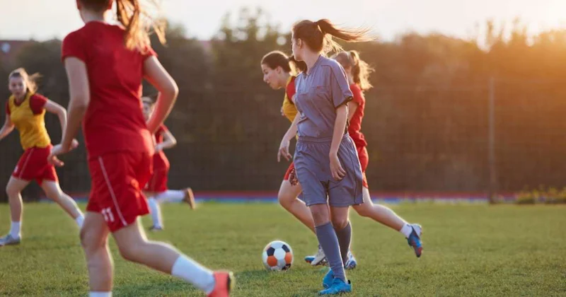 Kırmızı formalar giyen bir grup kız çimenli bir sahada futbol oynuyor. Görüntünün odağında ön planda, sırtı kameraya dönük, elinde topla koşan bir kız var. Kırmızı tişörtlü bir başka kız da onun yanında koşuyor. Görüntünün arka planı bulanık, ancak uzakta koşan ve oynayan kırmızı üniformalı iki kız daha görülebiliyor. Çimlerin üzerinde duran bir çift ayağın yakın çekimi de görülebiliyor. Çimler yemyeşil ve gür ve çimlerin üzerinde bir futbol topu görülebiliyor.