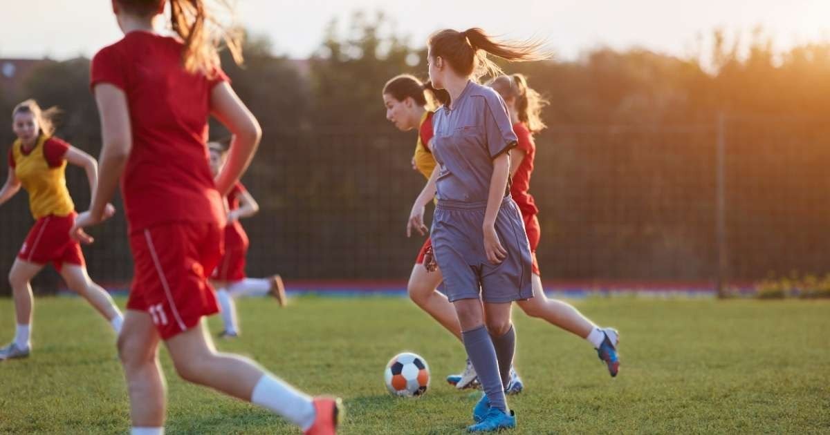 Kırmızı formalar giyen bir grup kız çimenli bir sahada futbol oynuyor. Görüntünün odağında ön planda, sırtı kameraya dönük, elinde topla koşan bir kız var. Kırmızı tişörtlü bir başka kız da onun yanında koşuyor. Görüntünün arka planı bulanık, ancak uzakta koşan ve oynayan kırmızı üniformalı iki kız daha görülebiliyor. Çimlerin üzerinde duran bir çift ayağın yakın çekimi de görülebiliyor. Çimler yemyeşil ve gür ve çimlerin üzerinde bir futbol topu görülebiliyor.