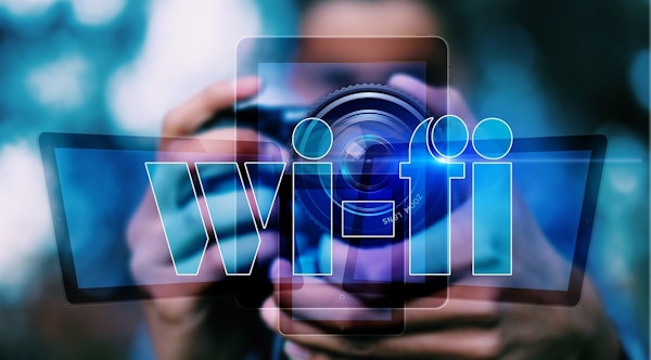 Bir kişi önünde bir dijital fotoğraf makinesi tutmaktadır. Fotoğraf makinesinin yan tarafında mavi beyaz bir logo vardır ve objektif çerçevenin ortasındadır, yakın plan bir görüntüye sahiptir. Kişi işaret parmağını deklanşör düğmesine koymuş, fotoğraf çekmeye hazır. Siyah bir gömlek ve kot pantolon giymekte, kahverengi bir kemer ve kahverengi bir sırt çantası taşımaktadır. Arka plan açık mavi bir duvar ve çerçevenin sol üst köşesinde bir pencere görülüyor. Arka planda üzerinde çeşitli eşyalar bulunan beyaz bir raf var ve odada birkaç bitki var.
