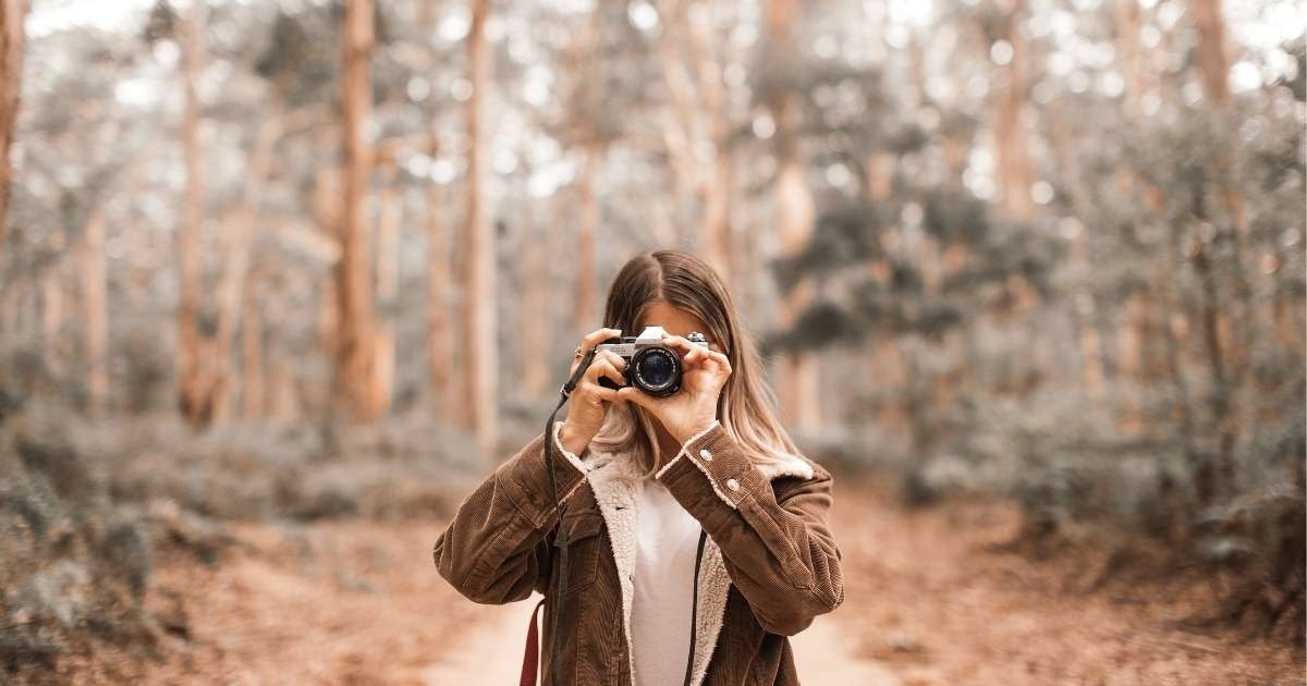 Siyah ceketli bir kadın iki eliyle yüzünün önünde bir dijital fotoğraf makinesi tutuyor. Hafifçe gülümseyen bir ifadeyle doğrudan objektife bakıyor, gözleri hafifçe kısılıyor. Kaşları çatılmış ve dudakları hafifçe ayrılmış. Kahverengi saçları arkaya toplanmış ve ceketinin altına beyaz bir tişört giymiş. Fotoğraf makinesi, vizörü, zoom lensi, flaşı ve bej renkli tutma yeri olan siyah dikdörtgen bir şekle sahip. Arka plan odak dışı yeşil ve kahverengi bir bulanıklık. Kendi fotoğrafını çekiyor gibi görünüyor.