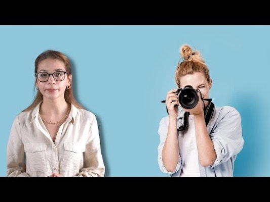 Koyu renk saçlı, gözlüklü ve beyaz gömlekli bir kadın bir elinde fotoğraf makinesi tutuyor. Parlak mavi bir arka planın önünde duruyor, diğer kolu avuç içi yukarı bakacak şekilde bükülmüş. Yüzünde kararlı bir ifade var, gözleri elindeki kameraya odaklanmış. Vücudu kameraya doğru hafifçe eğilmiş, duruşu kendinden emin ve iddialı. Bir fotoğraf çekme sürecindeymiş gibi görünüyor.
