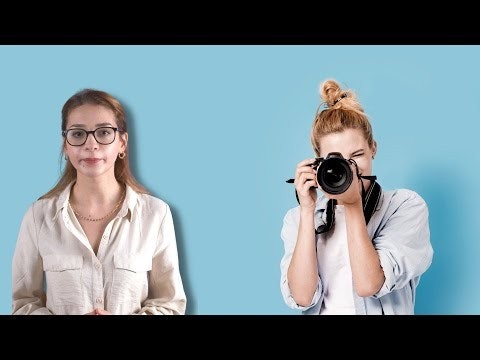 Koyu renk saçlı, gözlüklü ve beyaz gömlekli bir kadın bir elinde fotoğraf makinesi tutuyor. Parlak mavi bir arka planın önünde duruyor, diğer kolu avuç içi yukarı bakacak şekilde bükülmüş. Yüzünde kararlı bir ifade var, gözleri elindeki kameraya odaklanmış. Vücudu kameraya doğru hafifçe eğilmiş, duruşu kendinden emin ve iddialı. Bir fotoğraf çekme sürecindeymiş gibi görünüyor.