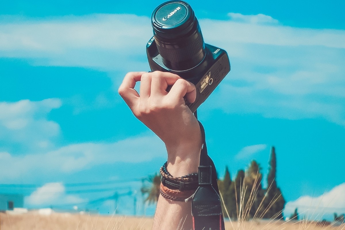 Resimde, elinde dijital bir fotoğraf makinesi tutan bir elin yakın çekimi görülüyor. Fotoğraf makinesinin gümüş detayları olan siyah bir gövdesi ve büyük bir lensi var. El, görüntüde odakta olan gümüş bir bilezik takıyor. Arka plan bulanıktır ve alt köşede küçük bir ağaç görünmektedir. Görüntü, fotoğraf makinesinin ve bileziğin her parçasının görülebildiği yüksek bir ayrıntı seviyesine sahiptir. Fotoğraf makinesinin lensi de odakta ve el yakın açıdan görülüyor. Görüntüdeki renkler çoğunlukla siyah ve gümüş tonlarında olup birkaç yeşil ipucu da içeriyor.