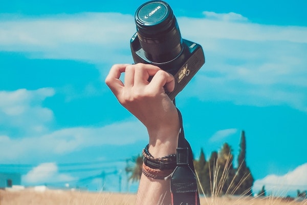 Resimde, elinde dijital bir fotoğraf makinesi tutan bir elin yakın çekimi görülüyor. Fotoğraf makinesinin gümüş detayları olan siyah bir gövdesi ve büyük bir lensi var. El, görüntüde odakta olan gümüş bir bilezik takıyor. Arka plan bulanıktır ve alt köşede küçük bir ağaç görünmektedir. Görüntü, fotoğraf makinesinin ve bileziğin her parçasının görülebildiği yüksek bir ayrıntı seviyesine sahiptir. Fotoğraf makinesinin lensi de odakta ve el yakın açıdan görülüyor. Görüntüdeki renkler çoğunlukla siyah ve gümüş tonlarında olup birkaç yeşil ipucu da içeriyor.