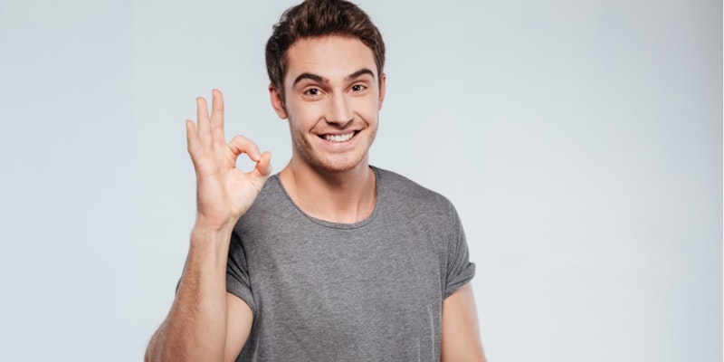 The man in the image is wearing a grey long-sleeved shirt, and his arms are bent in an upward direction with his hands near his face. His eyes are closed and his mouth is slightly open, as if he is making a sound. He is standing in front of a white background, with nothing else in the frame. His posture is relaxed and his expression is peaceful. He appears to be in a state of deep contemplation, perhaps lost in thought. His attire is casual and the color palette of his clothing is subtle. His face is illuminated by a natural light source, giving him a gentle and soothing look. He appears content and comfortable in his environment.
