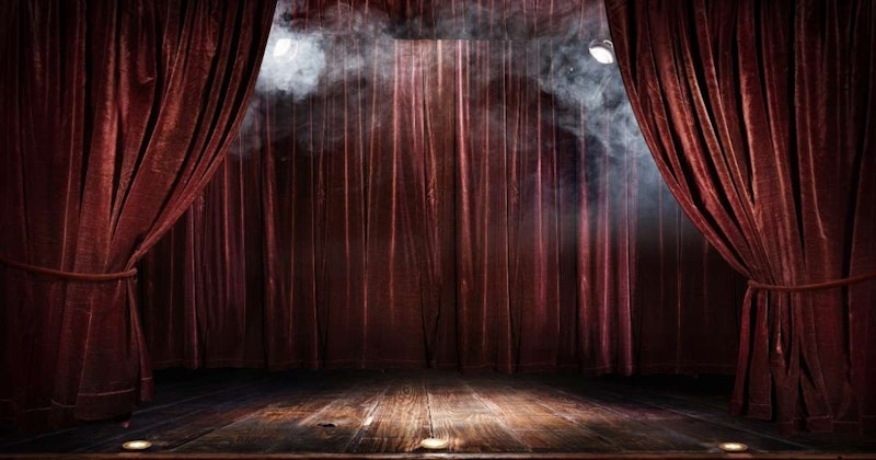 Bu fotoğrafta ahşap zeminli ve kırmızı perdeli bir sahne görülüyor. Zemin orta tonlu bir ahşap renginde ve birkaç küçük eşya ile kaplı. Perdeler koyu, canlı bir kırmızıdır ve iki kat halinde asılıdır. Üst katman alt katmandan biraz daha açık renktedir. Perdeler ortada toplanmış ve yere kadar sarkıyor. Ahşap zeminde parlayan bir ışık ve sahnenin sol tarafından yükselen az miktarda duman vardır. Perdeler hafifçe geriye çekilerek alttaki ahşap zeminin görünmesini sağlıyor. Fotoğrafın kompozisyonu dengeli ve perdeler ile zeminin renkleri güzel bir kontrast oluşturuyor. Fotoğraf sahnenin yakın çekimidir ve izleyiciye güzel ortamın ayrıntılarına samimi bir bakış sağlar.