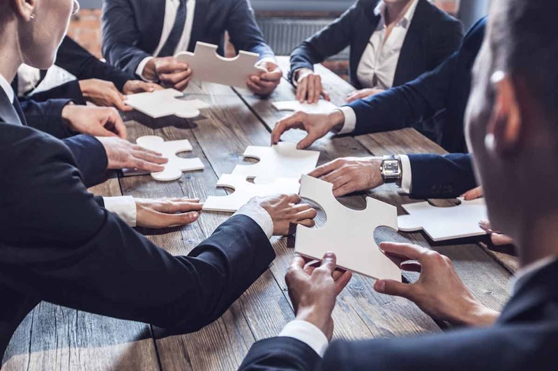 Hepsi de çeşitli kıyafetler giyen beş kişilik bir grup, bir masanın etrafında oturmaktadır. Bir kadın mavi bir elbise giyerken, bir diğeri yeşil bir gömlek giymektedir. Başka bir kadın siyah beyaz çizgili bir gömlek ve kot pantolon giymektedir. Dördüncü kişi siyah gömlek ve kot pantolon giyen bir erkek. Beşinci kişi kolunda beyaz ve mavi bir saat ve beyaz ve mavi bir gömlek giyen bir adamdır. Tüm insanlar ellerinde yapboz parçaları tutmakta ve onlara dikkatle bakmaktadır. Grubun yakın plan çekimi olduğundan her bir kişinin yüz ifadesini ve yapboz parçalarının detaylarını görebiliyorsunuz.