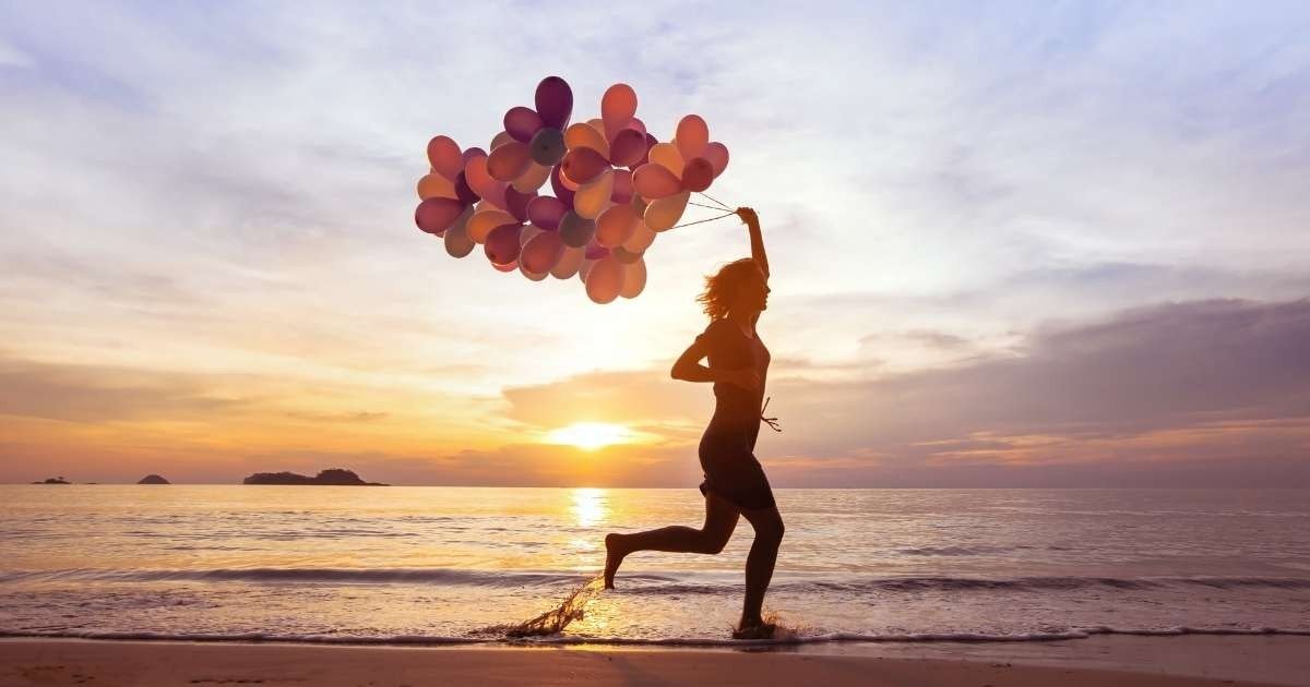 Arka planda canlı bir gün batımı ile sahilde koşan bir kadın, elinde bir demet renkli balon taşıyor. Hareket ettikçe saçları arkasında uçuşuyor ve arka planda deniz şırıldıyor. Dalgalar kıyıya vuruyor ve ışık sudan yansıyarak sakinleştirici bir atmosfer yaratıyor. Kadının silueti gökyüzünün turuncu ve pembe tonlarında göze çarpıyor ve elinde tuttuğu balonlar parlak bir şekilde aydınlatılıyor. Kadın, balonlarla koşarken ve çevresinin güzelliğini seyrederken neşeli bir an yaşıyor gibi görünüyor.