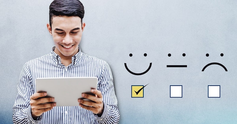 Parlak bir gülümsemeye sahip bir adam oturmuş, elindeki siyah tablete dikkatle bakıyor. Koyu renk saçları var ve gözleri kapalı, bu da ona memnun bir ifade veriyor. Tabletin üzerinde, içinde siyah bir onay işareti olan sarı bir kare var. Arka planda siyah çizgilerden oluşan beyaz bir fon ve mavi kenarlıklı beyaz bir kare var. Adam görüntüdeki tek kişidir ve çerçevenin ortasındadır. Gri bir gömlek giyiyor ve odağı elindeki tablette.