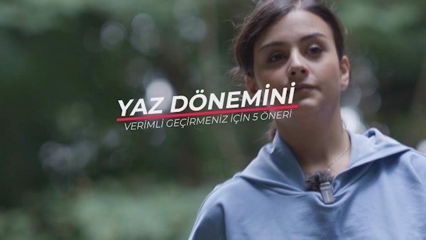 Mavi sweatshirt giyen bir kadın parlak beyaz bir arka planın önünde duruyor. Kolları iki yanında, yüzü kameraya dönüktür. Saçları yüzünden geriye doğru toplanmıştır ve yüz hatları açıkça görülebilmektedir. Lacivert bir kapüşonlu sweatshirt giymektedir ve fermuarı önden aşağı doğru inmektedir. Kapüşonlu sweatshirt'ün kapüşonu yukarı çekilmiş ve fermuarı yarı açık. Sweatshirt'ün önündeki logo açıkça görülebiliyor. Çerçevenin sağ alt köşesinde beyaz zemin üzerine siyah harflerle yazılmış bir tabela var. Harflerde Hoş geldiniz yazmaktadır.