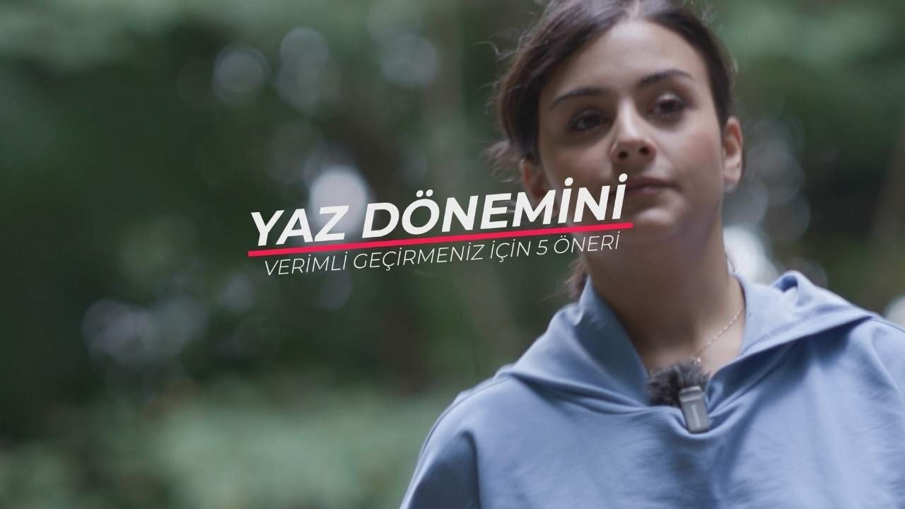 Mavi sweatshirt giyen bir kadın parlak beyaz bir arka planın önünde duruyor. Kolları iki yanında, yüzü kameraya dönüktür. Saçları yüzünden geriye doğru toplanmıştır ve yüz hatları açıkça görülebilmektedir. Lacivert bir kapüşonlu sweatshirt giymektedir ve fermuarı önden aşağı doğru inmektedir. Kapüşonlu sweatshirt'ün kapüşonu yukarı çekilmiş ve fermuarı yarı açık. Sweatshirt'ün önündeki logo açıkça görülebiliyor. Çerçevenin sağ alt köşesinde beyaz zemin üzerine siyah harflerle yazılmış bir tabela var. Harflerde Hoş geldiniz yazmaktadır.