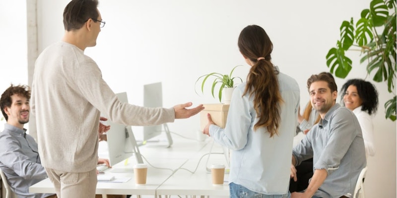 Uzun, siyah saçlı bir kadın, içinde beyaz bir saksı olan bir kutunun önünde duruyor. Gri bir gömlek giymiş ve gözlük takmış olan kadın kameraya gülümsüyor. Kutu elleriyle vücudunun önünde tutulmaktadır. Arkasında bir adam bilgisayarın önünde durmaktadır ve sağında yüzü bulanık olan başka bir kişi vardır. Arka plan karanlık ve görüntü odaklanmış. Kadının yüz ifadesi neşe ve memnuniyet dolu.