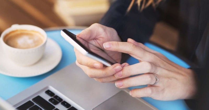 Bir kişi elinde kameraya dönük bir akıllı telefon tutmaktadır. Telefonun siyah bir kasası vardır ve kameranın ekranı yakalamasını sağlayacak şekilde tutulmaktadır. Kişi açık renkli bir bluz giyiyor ve sol elinde bir kol saati var. Görüntü, kişinin ellerine ve telefona odaklanan bir yakın çekimdir. Arka plan odak dışıdır ve nötr bir renkte görünmektedir. Kişinin elleri ön plandadır, parmaklarındaki ve telefondaki ayrıntılar görülmektedir. Görüntü hafif sarımsı bir renk tonuna sahiptir ve hafif sıcak bir atmosfer verir. Telefonun ekranı da görülebiliyor ve bir klavye görüntüsü gösteriyor.