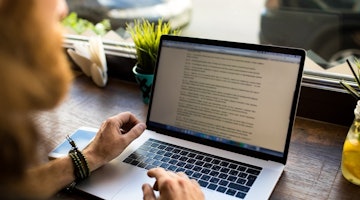 Bir kişi masa başında oturmuş dizüstü bilgisayarında bir şeyler yazıyor. Dizüstü bilgisayar siyah ve gümüş renklidir ve kişinin her iki bileğinde de bilezikler vardır ve bunlardan biri dizüstü bilgisayarın klavyesinde görünmektedir. Arka planda üzerinde siyah bir sembol olan mavi bir fincan var. Kişi ekrana odaklanmış, sağ eliyle klavyedeki bir düğmeye basıyor. Görüntü yakın çekimdir ve dizüstü bilgisayarın tuşları, fincan ve sembolü ile bilezikler hakkında ayrıntılı bilgi vermektedir. Kişi gözlük ve mavi bir gömlek giymektedir.