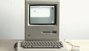 Bu görüntü klavyeli eski bir bilgisayara aittir ve klavyenin yakın çekimini içermektedir. Bilgisayarın sol üst köşesinde beyaz bir kutu bulunan bir ekran ve sağ alt köşesinde bir fare yer almaktadır. Ayrıca, sol alt köşede bir tel ve bir spiralin yakın çekimi görülebilmektedir. Klavye eski bir model gibi görünmektedir ve tuşları hafif kavisli bir şekle sahiptir. Fare, üst yüzeyinde iki düğme bulunan küçük, kablolu bir versiyon gibi görünmektedir. Bilgisayar ekranındaki beyaz kutu hafifçe yükseltilmiş ve ince çizgilerden oluşan bir desene sahiptir.