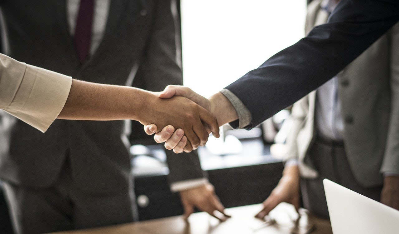 Two people wearing formal attire stand side by side and shake hands in a close-up shot. The man on the left is wearing a light grey suit with a white shirt, a tie, and a pair of black leather shoes. His hand is gripping the other person's hand tightly. The man on the right is wearing a navy blue suit with a light blue shirt, a black tie, and a pair of brown leather shoes. His hand is clasped around the other person's in a firm handshake. Both individuals have a look of determination and seriousness on their faces. Their hands are slightly blurred in the background, suggesting a swift and vigorous motion.