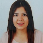 Omuz hizasında kumral saçları olan bir kadın yüzünde kocaman, ışıl ışıl bir gülümsemeyle ayakta duruyor. Üzerinde beyaz yakalı ve çiçek desenli pembe bir bluz var ve saçları rüzgârda hafifçe dalgalanıyor. Dudakları neşeli bir ifadeyle kıvrılmış, gözleri parlak ve mutlu. Ellerini kalçalarına koymuş ve soluk mavi bir arka planın önünde duruyor. Uzun bukleleri sırtından aşağı dökülüyor ve ışıkta parlıyor. Memnun ve neşeli görünüyor, güven ve pozitiflik duygusu yayıyor.