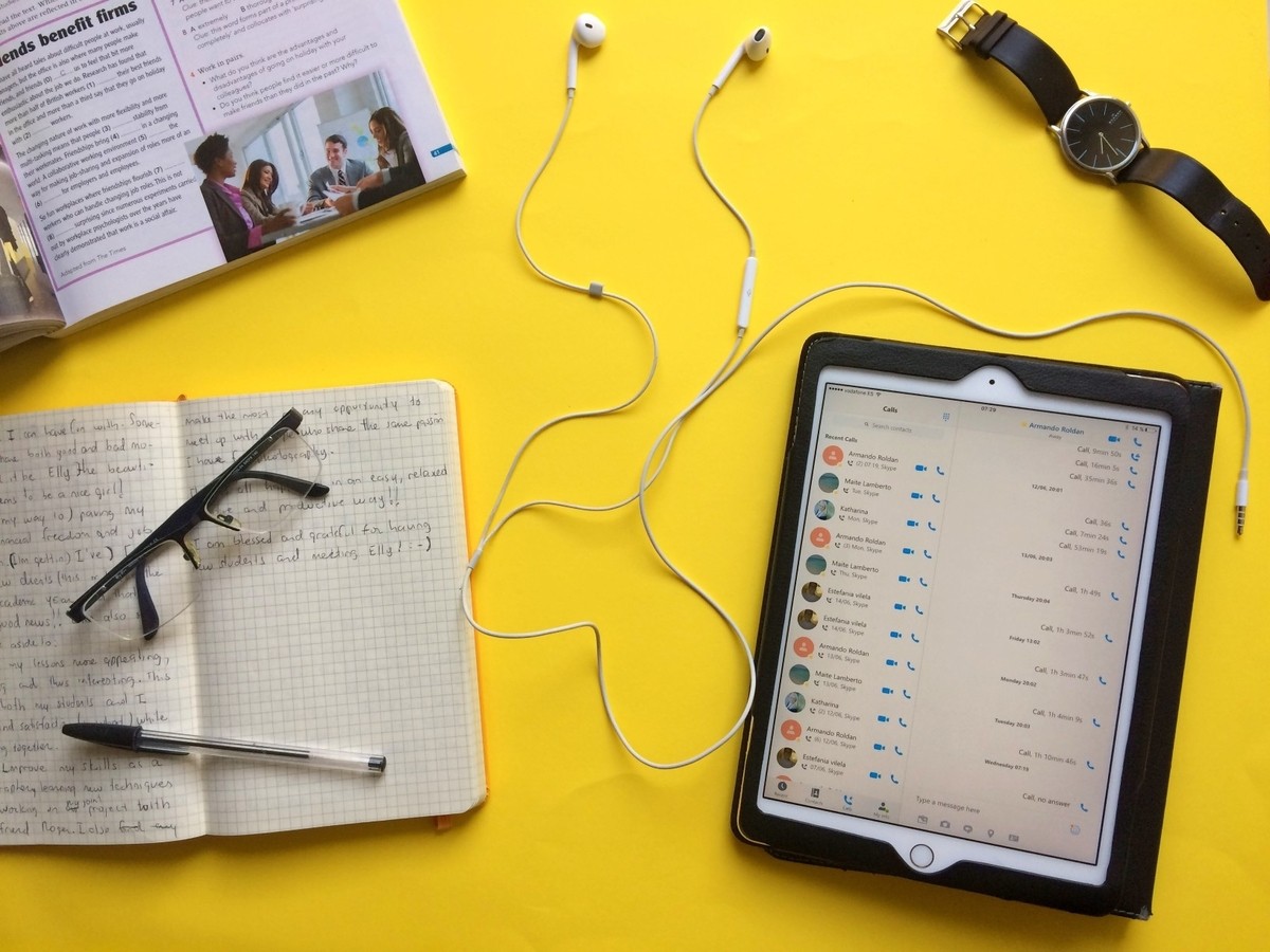 Sarı arka planda bir tablet ve bir çift kulaklık görülüyor. Tabletin solunda bir not defteri, üzerinde bir kalem ve gözlük yer alıyor. Tabletin sağında deri kayışlı bir saat var. Arka planda, karo bir duvarın üzerinde metal bir çubuk var. Görüntünün ortasında bir kadının yakın çekimi ve bir kağıt parçasının yakın çekimi görülüyor. Tabletin aydınlatılmış bir ekranı vardır ve kağıt üzerinde metin ve görüntüler görülebilmektedir. Görüntüdeki öğelerin renkleri canlıdır ve sarı arka plan büyük bir kontrast sağlamaktadır. Genel olarak, bu ilginç bir bağlam sağlayan ilginç bir görüntüdür.