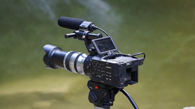 Üzerine kamera monte edilmiş bir tripod bu görüntünün odak noktasıdır. Kamera çerçevenin ortasına yerleştirilmiş ve alt kısmında kavisli bir metal nesne var. Kamera merceği yakın plandadır ve yan tarafında mavi bir nesne vardır. Kamera kafası da çerçevede görülebiliyor ve yanında siyah bir video kaydedici var. Objektifin net bir görüntüsü odaktadır ve kamera ile tripodun renkleri iyi tanımlanmıştır. Görüntünün tüm ayrıntıları çok nettir, bu da onu bir görüntü başlığı veri seti hazırlamak için ideal kılar.