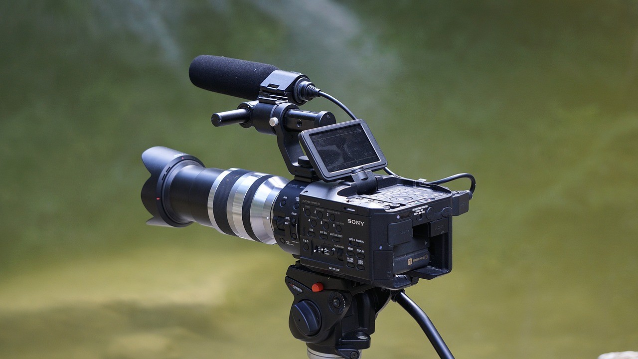 Üzerine kamera monte edilmiş bir tripod bu görüntünün odak noktasıdır. Kamera çerçevenin ortasına yerleştirilmiş ve alt kısmında kavisli bir metal nesne var. Kamera merceği yakın plandadır ve yan tarafında mavi bir nesne vardır. Kamera kafası da çerçevede görülebiliyor ve yanında siyah bir video kaydedici var. Objektifin net bir görüntüsü odaktadır ve kamera ile tripodun renkleri iyi tanımlanmıştır. Görüntünün tüm ayrıntıları çok nettir, bu da onu bir görüntü başlığı veri seti hazırlamak için ideal kılar.