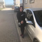 Kadın mavi bir arabanın yanında duruyor. Beyaz bir üst ve mavi bir kot pantolon giyiyor. Saçları uzun ve kahverengidir. Ciddi bir ifadeyle kameradan uzağa bakmaktadır. Kollarını önünde kavuşturmuş. Araba krom aksanlı ve alaşım jantlı dört kapılı bir sedan. Arka planda ağaçların olduğu asfalt bir yola park edilmiş. Güneş parlak bir şekilde parlıyor ve yerde gölgeler yaratıyor.