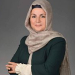 Saçları düzgünce arkadan bağlanmış genç bir kadın parlak mavi bir başörtüsü takıyor. Cildi sıcak, karamel renginde ve gözleri koyu kahverengi. Kameraya bakarken yüzünde nazik, gülümseyen bir ifade var. Beyaz bir üst ve mavi ve yeşil çiçek desenli, desenli, uzun bir etek giyiyor. Bir çift küpe ve altın bir kolye ile aksesuarlarını tamamlamış. Kıyafetleri, içinde bulunduğu güneşli, açık hava ortamına mükemmel bir uyum sağlıyor.