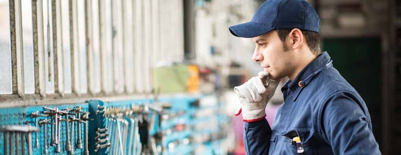 Mavi şapkalı ve eldivenli bir adam bir duvarın önünde durmuş, yerdeki bir alet rafına bakmaktadır. Gümüş kenarlı mavi bir şapka takıyor ve eldivenleri beyaz ve gri desenli. Sağ tarafta bir sıra mavi ve gümüş sandalye var. Ön planda, bir eldiven ve bir aletin yakın çekimi görülebiliyor, ikisi de odak dışı. Adamın mavi gömleği de arka planda görülebiliyor. Beyaz duvarlı ve karo zeminli bir odada durmaktadır.