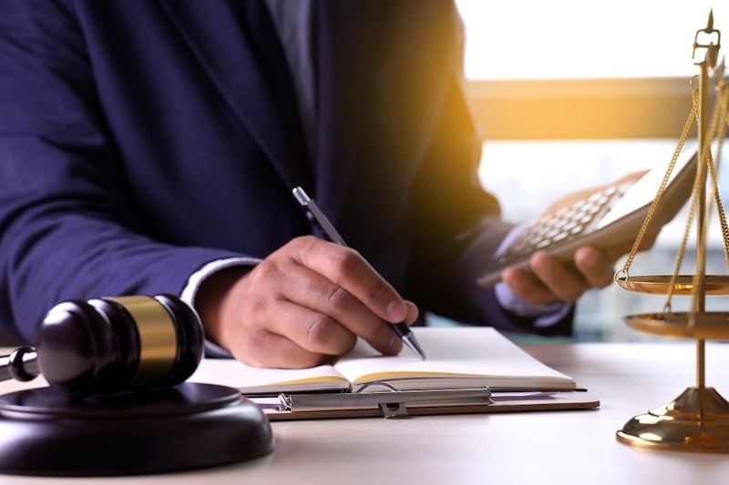 Bir kişi kalem ve hesap makinesi tutuyor. Görüntü, arka planda siyah ve altın rengi bir nesne ile kişinin elinin yakın çekimidir. El kalemi sıkıca kavramaktadır ve hesap makinesi diğer elinde görülebilmektedir. Kişi bir deftere yazıyor ve görüntünün köşesinde bir ağırlık ölçeği görülebiliyor. Her şey hafif bulanık, bu da resme rüya gibi bir his veriyor. Kişi elindeki işe odaklanmıştır ve ifadesi yoğun ve kararlıdır.