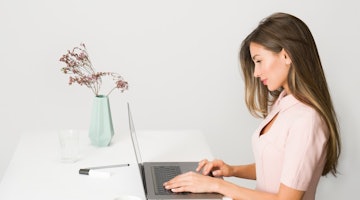 Bir kadın masada oturmuş dizüstü bilgisayar kullanıyor. Üzerinde pembe bir elbise var ve eli dizüstü bilgisayarın klavyesinde. Önündeki masada bir fincan kahve ve bir lokum var. Solunda yeşil saplı beyaz bir nesne ve yanında pipetli bir bardak vardır. Arkasında içinde çiçekler olan bir vazo ve sağında beyaz bir yüzey üzerinde metal bir çubuk var.