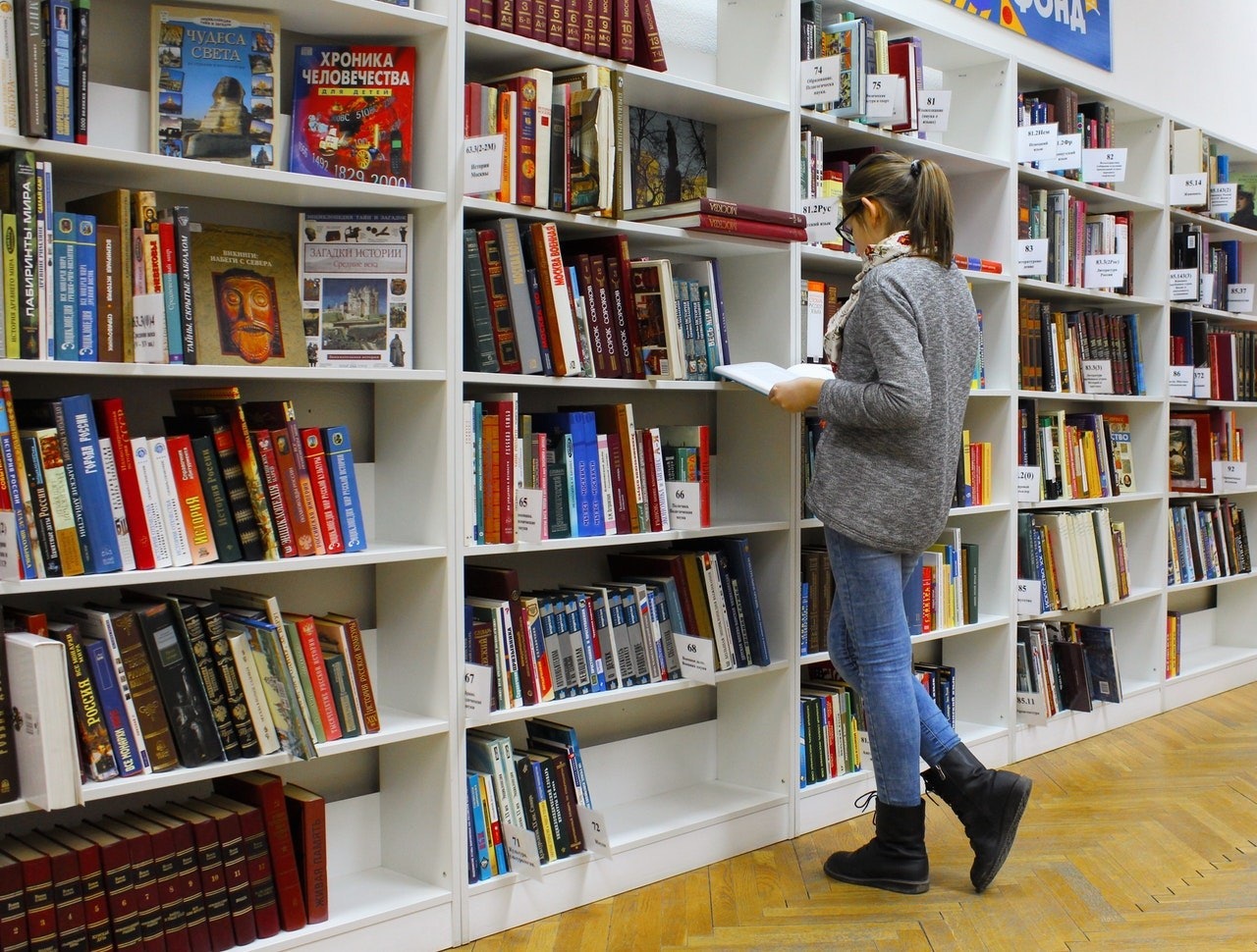 Gri kazaklı bir kadın bir kitap rafının önünde durmuş, kitaplara bakmaktadır. Rafın sol tarafında, üzerinde telefon resmi olan bir kitap ve üzerinde kale resimleri olan bir poster var. Sağ tarafta, üzerinde resimler olan bir kitap ve üzerinde bir yüz olan bir kitap var. Rafın alt kısmında bir çift siyah çizme görülüyor. Kadının sağında, üzerinde siyah yazılar olan beyaz bir tabela var.