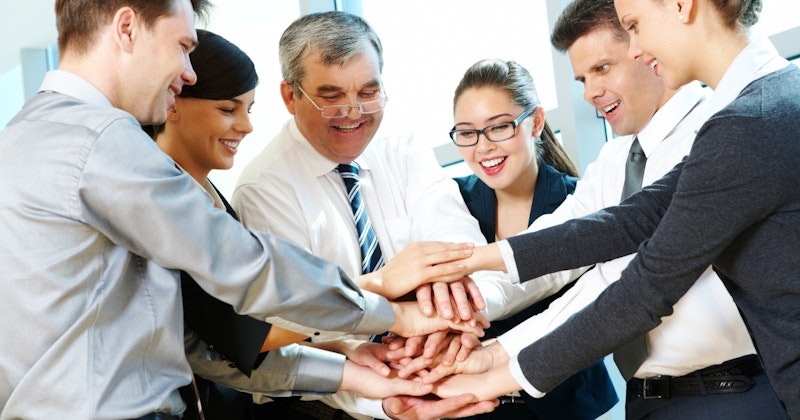 Gülümseyen ve mutlu bir grup insan, ellerini birleştirmiş bir daire içinde durmaktadır. Gruptaki bireylerden ikisi, bir erkek ve bir kadın, birbirlerini kucaklıyorlar. Adamın üzerinde mavi bir takım elbise ve siyah bir kemer var. Kadın gözlük takıyor ve gülümsüyor. Arka planda başka bir kişinin yüzünün yakın çekimi ve bir kravatın yakın çekimi var. Gruptaki tüm insanlar gözlerini kapatmış, neşe ve birlik içindedirler.