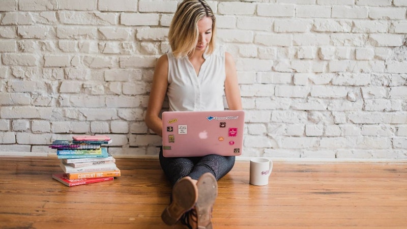 Genç bir kadın yerde oturmuş, bir çift beyaz spor ayakkabı giyiyor. Sarı saçları var ve kucağında duran dizüstü bilgisayarına dalmış durumda. Önündeki masada bir yığın kitap ve yanında pembe logolu beyaz bir kupa var. Ayakları yere basmakta ve bir şeyler yazarken kolları tembelce önünde uzanmaktadır. Dizüstü bilgisayar ekranı net bir şekilde görülebiliyor ve klavye yumuşak bir ışıkla aydınlatılıyor. Tamamen işine odaklanmış durumda ve sahne huzurlu ve dingin.