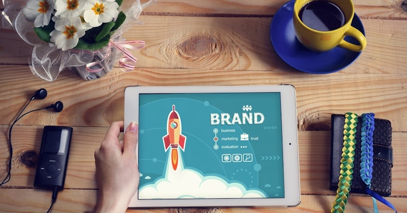 Tablet cihaz tutan bir elin yakın plan çekimi ve ekranda gökyüzüne fırlatılan parlak sarı bir roket. Tablet yumruk benzeri bir tutuşla tutulmakta ve roket, alt kısmında parlak turuncu ve sarı bir alevle tam olarak gösterilmektedir. Arka plan, dağınık beyaz bulutlarla koyu mavi bir gökyüzüdür. Çerçevenin sol tarafında içi sıvı dolu sarı bir kupa, sağ tarafında ise saksı içinde beyaz bir çiçek yer almaktadır. Daha sağda ise mavi bir tabağı olan sarı bir fincan yer alıyor. Görüntü keskin ve net, renkler ise canlı ve parlak. Roket güçlü bir patlamayla havalanıyor gibi görünürken, ön plandaki el bir ölçek duygusu katıyor.