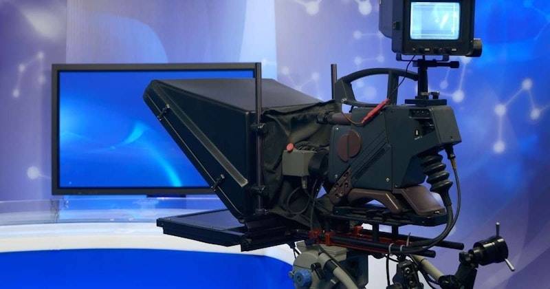 Bu görüntü bir stand üzerindeki kameranın yakın çekimini göstermektedir. Kamera siyahtır, mavi bir ekranı ve ortasında beyaz bir çarpı işareti vardır. Kameranın sağında küçük kare şeklinde siyah bir cihaz var. Ön planda, gri ve siyah bir kasaya ve siyah bir kontrol paneline sahip bir makinenin yakın çekimi var. Daha geride, siyah ve gri gövdeli daha büyük bir makine vardır. Sahne, kamera ve makinelerdeki vurgularla iyi aydınlatılmıştır.