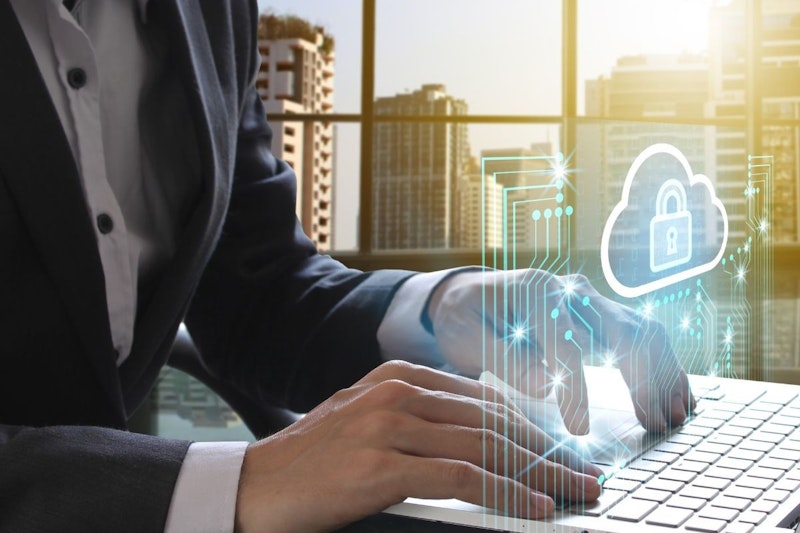 Bir kişi, elleri ve parmakları klavyenin üzerinde konumlanmış şekilde dizüstü bilgisayarda yazı yazıyor. Elleri hafifçe birbirinden ayrılmış ve gözleri ekrana odaklanmıştır. Sağ üst köşede kilit simgesi olan bir bulut görülüyor. Dizüstü bilgisayar ekranı hafif bulanık, bu da kişinin bir süredir yazdığını gösteriyor. Görüntü yakın çekim hissi veriyor ve kişinin kolu sağ alt köşede görülebiliyor. Sahne soluk, beyaz bir arka planla çevrilidir. Kişinin vücut dili odaklanmış ve niyetlidir, bu da aktif olarak görevle meşgul olduğunu göstermektedir.