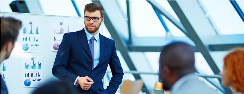 Siyah takım elbise giymiş ve kravat takmış bir adam beyaz tahtanın önünde duruyor. Koyu renk sakalı var ve yana doğru bakıyor. Yüz hatları biraz bulanık ama sağ kulağı net. Arkasındaki beyaz tahta grafikler ve diyagramlarla dolu. Görüntünün sol alt köşesinde küçük, bulanık beyaz bir nesne var. Adam tahtadaki bilgiler üzerine düşünüyor gibi görünüyor. Elleri cebinde, ciddi ve düşünceli görünüyor.