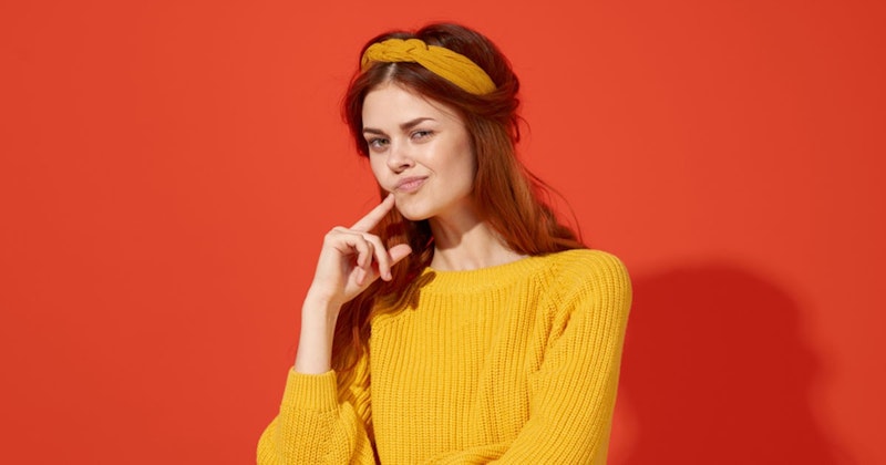 Parlak sarı bir kazak giyen genç bir kadın kameraya dönük duruyor. Omuz hizasında kahverengi saçları var ve yüzü sıcak, güneşli bir ışıkla aydınlanıyor. Sarı bir saç bandı takıyor ve ellerini önünde kavuşturmuş. Gözleri gökyüzüne doğru bakıyor ve yüzünde hafif bir gülümseme var. Sarı kazağı mavi gökyüzüne ve arka plandaki yeşil ağaçlara karşı öne çıkıyor ve görüntüye neşeli, canlı bir atmosfer veriyor. Kıyafetinin ve arka planın renkleri birlikte çalışarak mutluluk ve neşe hissi veren canlandırıcı bir görüntü oluşturuyor.