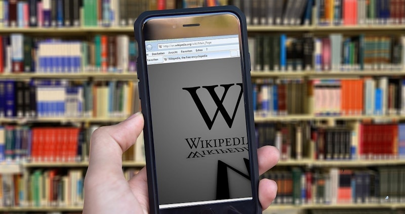 Parlak yüzeyli siyah bir cep telefonu tutan bir elin yakın çekimi. Telefon yatay bir pozisyonda tutulmakta ve kişinin parmakları kenarları sararken görülmektedir. Ekranda beyaz bir harf görülüyor ve arka planda soluk kırmızı ve beyaz damalı bir desen görülebiliyor. Ayrıca görüntünün köşesinde siyah çizgileri olan turuncu bir metal yüzey görülmektedir.