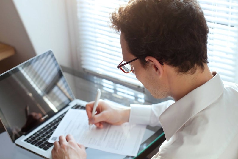 Sağ elinde tuttuğu kalemle bir kâğıt parçasına yazı yazan bir adamın yakın çekimi. Sol eli kâğıdı hafifçe kaldırılmış bir pozisyonda desteklemektedir. Kısa kahverengi saçları ve hafif bir sakalı var. Gözleri kâğıda odaklanmış ve kaşları konsantrasyonla hafifçe çatılmış. Yüzü sıcak sarı bir ışıkla aydınlatılmış. Kâğıt, yazdığı kelimeler dışında beyaz ve boş. Gömleği koyu mavidir ve kolları dirseklerine kadar kıvrılmıştır. Arka plan odak dışıdır ve sahneye atmosfer duygusu katan yumuşak bir bulanıklık vardır.